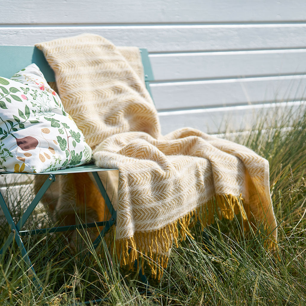 A cream and mustard yellow herringbone knit throw blanket with fringe is draped over a teal folding chair in a grassy outdoor setting next to a patterned pillow.