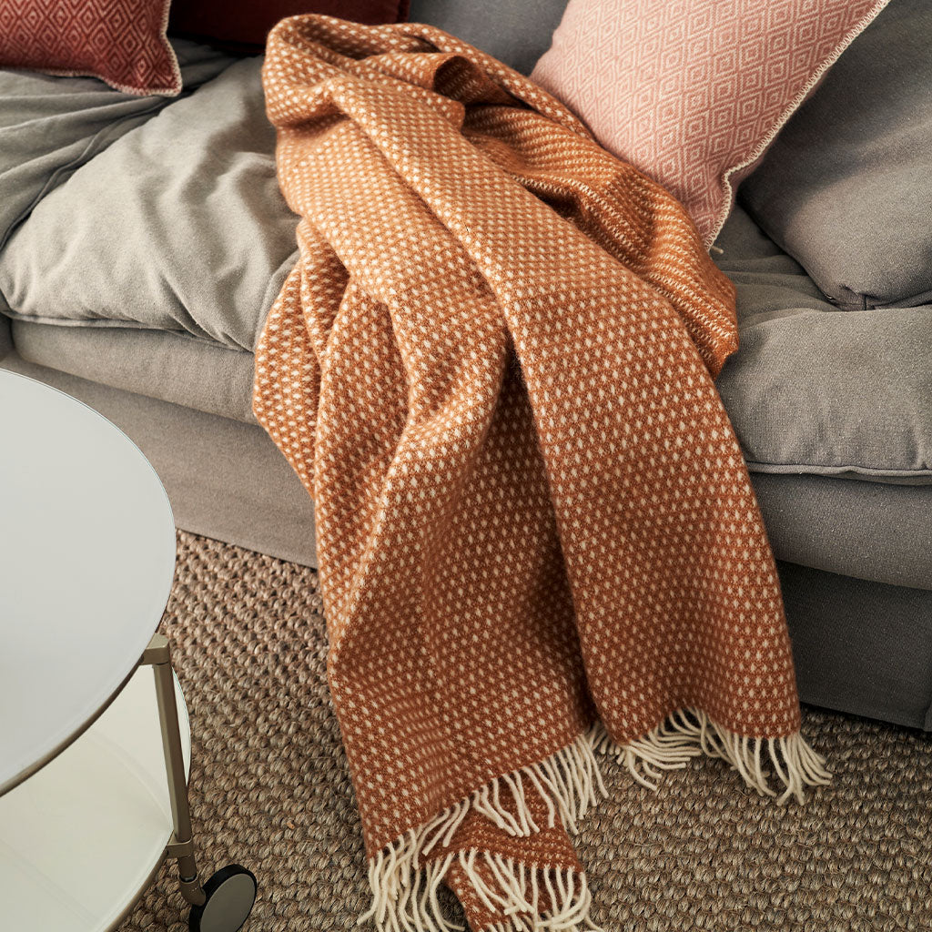 A rust-orange woven throw blanket with white fringe and a small white geometric pattern, draped over a gray sofa with pillows.