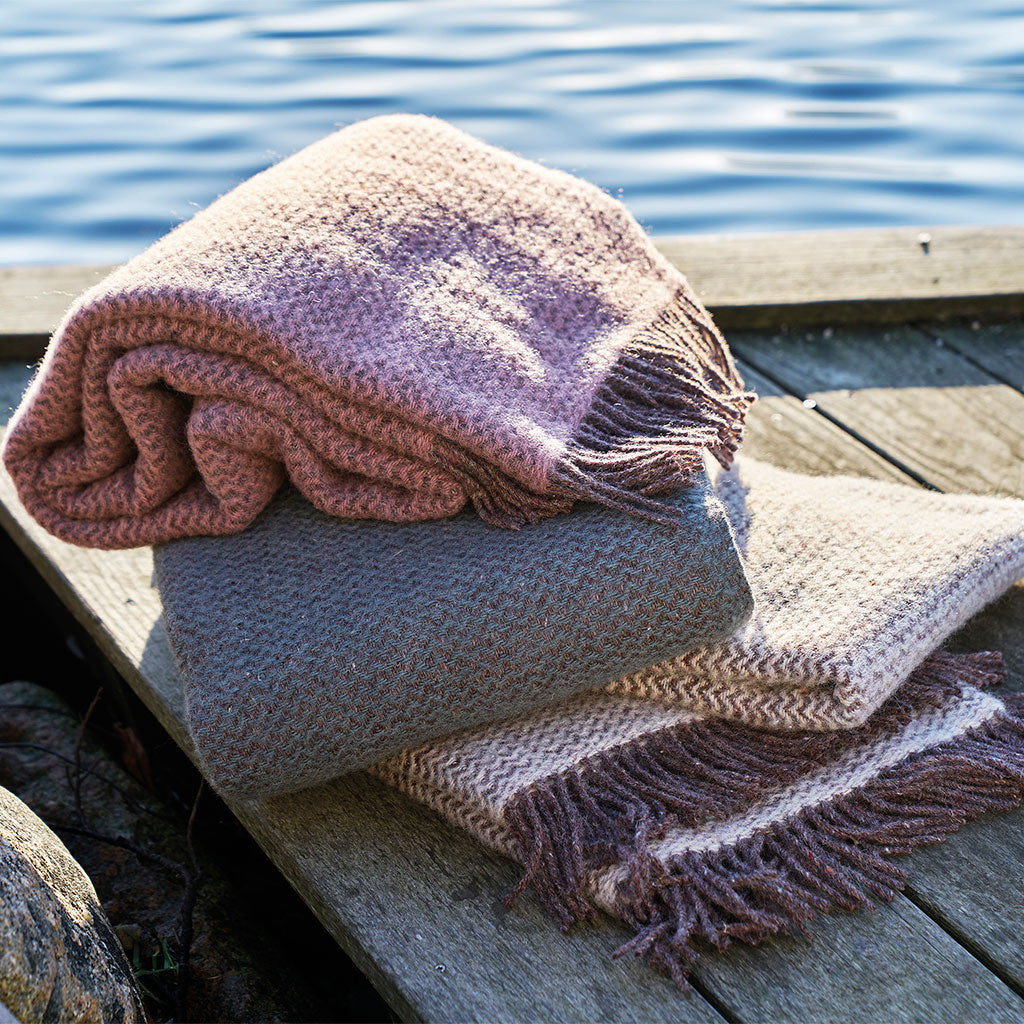 A folded knit throw blanket in dusty pink and taupe tones rests on a weathered wooden dock with blue water in the background.